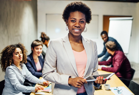 woman holding a tablet computer standing in front of a conference table of people while smiling at the camera