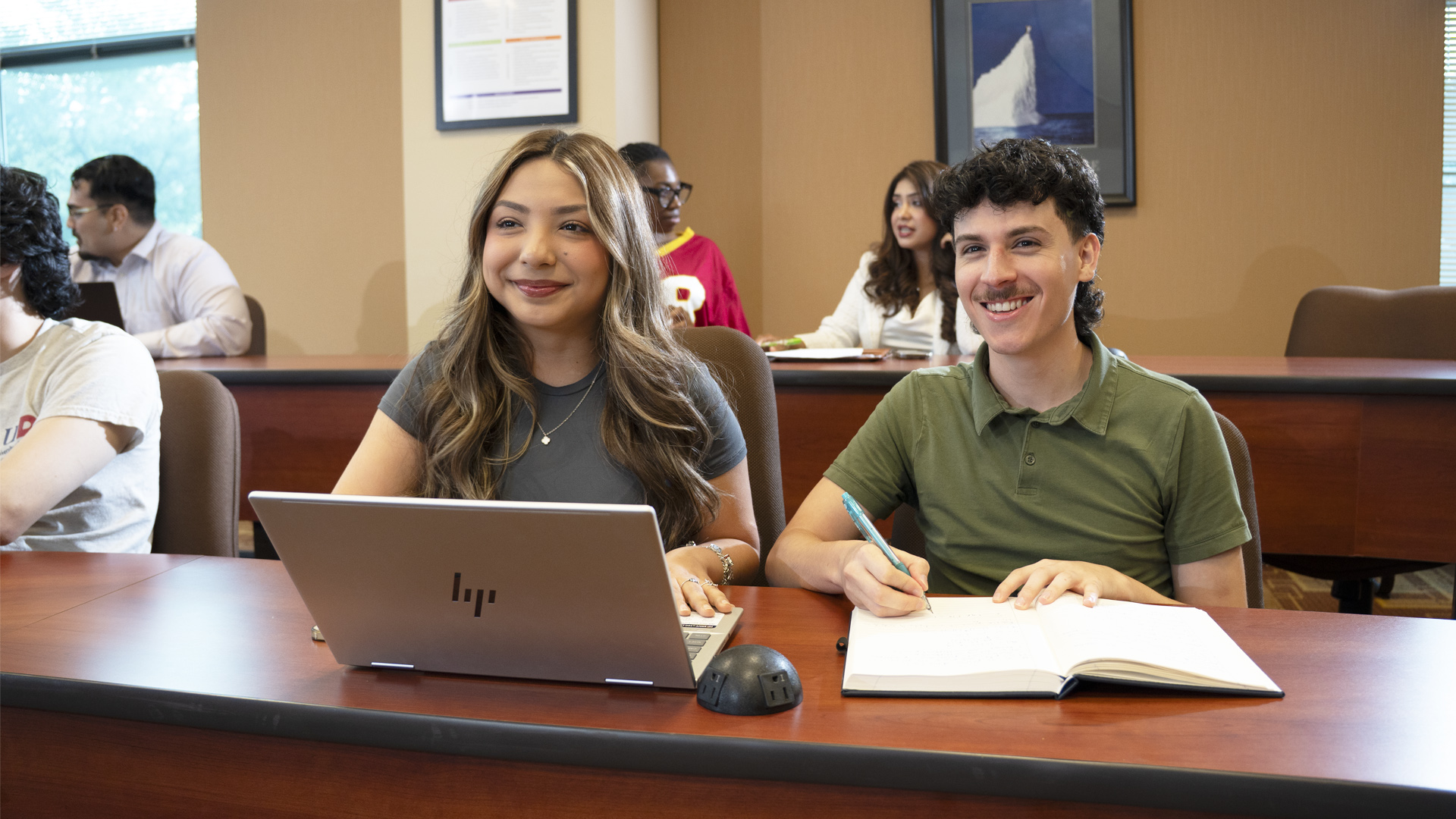 UHD female student sitting at a classroom desk with a laptop with a male UHD student sitting next to her with an open notebook ready to write notes