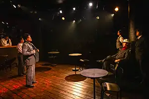 actor wearing sport coat faces two standing actors and one seated actor