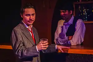 actor in pin striped cuts holds a drink in front of a bar tender