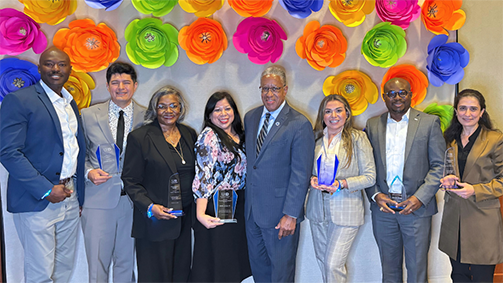 Seven adults hold glass awards while standing in front of a backdrop decorated with paper flowers at the university of houston-downtown alumni awards celebration, legacy and light.