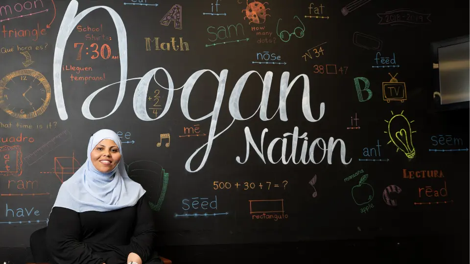 A woman in a hijab sits in front of a black wall featuring the words "Dogan Nation" prominently displayed