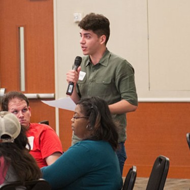 Students at round tables, one student standing with a mic in his hand