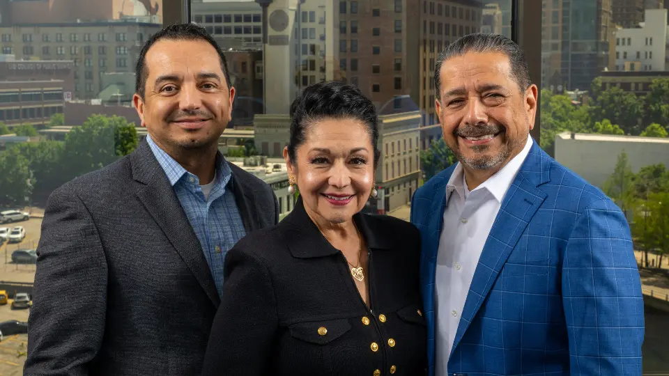 University of Houston-Downtown alums Robert Mendoza Jr, Paula Mendoza, and Robert Mendoza smile with the Downtown Houston skyline behind them in Houston, Texas.