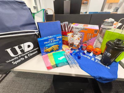 Snacks, a drawstring backpack, pens, a water bottle, a UHD Sports and Fitness drawstring backpack, and face masks on a table. The items are prizes for a gym membership drive for employees at the university of houston-downtown.