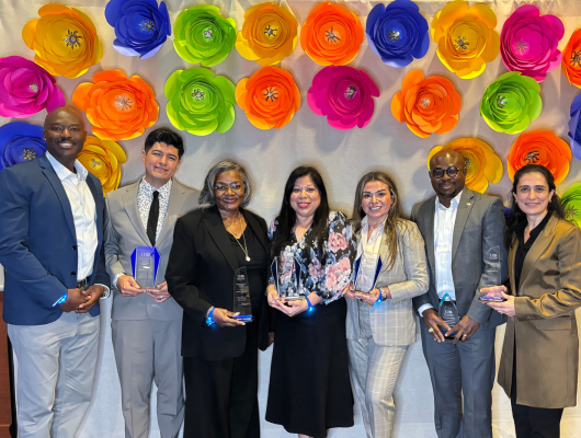 Seven adults hold glass awards while standing in front of a backdrop decorated with paper flowers at the university of houston-downtown alumni awards celebration, legacy and light.