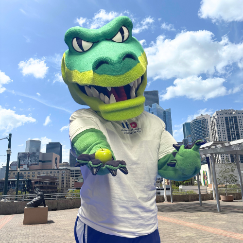 University of Houston-Downtown mascot Ed-U-Gator holds a green apple on the university's south deck with the Downtown Houston skyline behind them. 